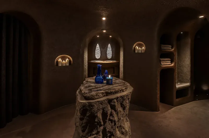 Cozy cave-like spa room with arched walls, tree stump table holding blue bottles, candles, towels on shelves