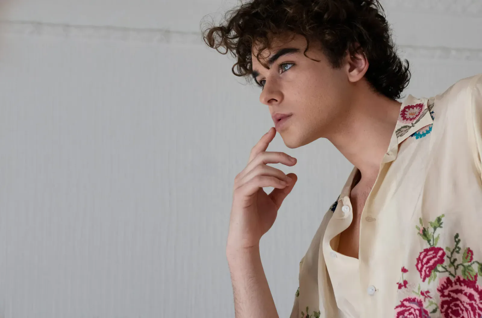 Javi Costa Polo in profile, curly hair, finger to lips, wearing open floral embroidered shirt against white background