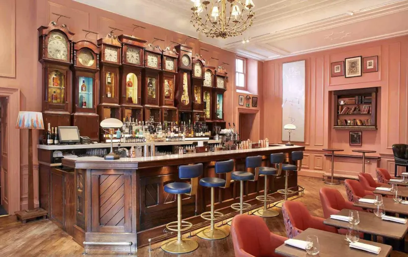 Elegant Home House bar in pink-walled room with wooden bar, blue stools, chandelier, antique clocks, and tables.