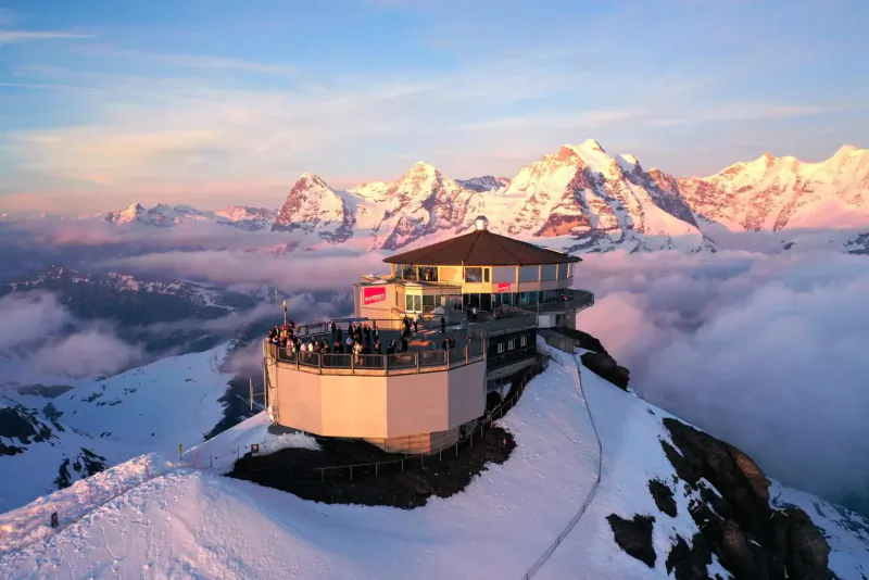 Mountain-top restaurant perched on snowy peak amid clouds, with Alps and sunset sky.