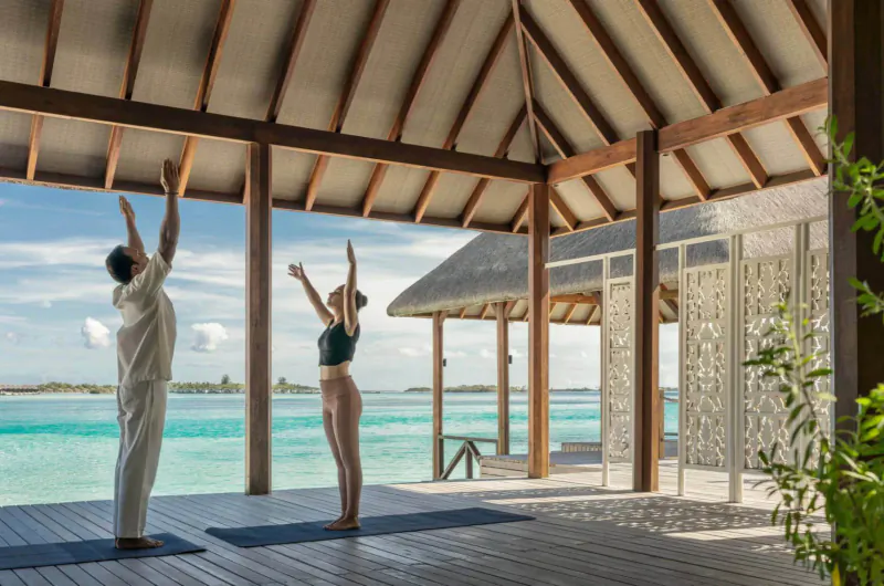 Man and woman in yoga poses with arms raised on mats in open wooden pavilion overlooking turquoise Maldives lagoon