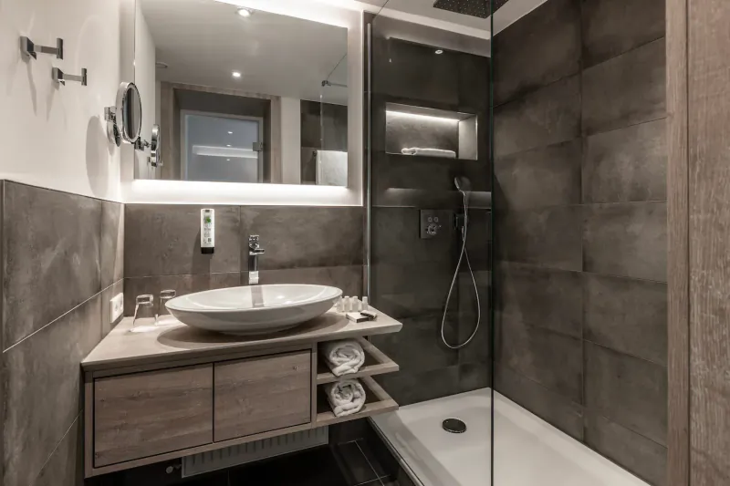 Modern hotel bathroom with wooden vanity, white bowl sink, rainfall shower, and gray stone tiles.