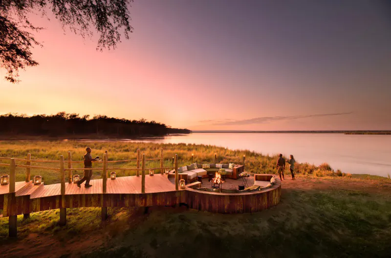 Sunset over wooden pier with fire pit and tables at Lavish Lolebezi luxury safari lodge in Zambia, two people walking.