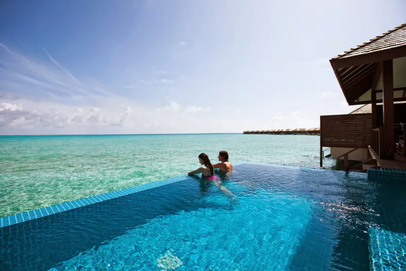 Couple relaxing in infinity pool at Hideaway Beach Resort, Maldives, with turquoise ocean and overwater villas beyond.