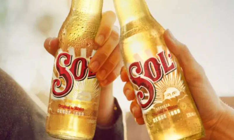 Two hands clinking chilled Sol cerveza bottles with condensation, red Sol labels and sun motifs.