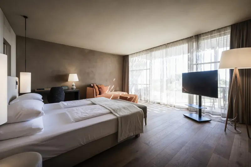 Spacious luxury hotel bedroom with king bed, orange armchair, TV, lamps, and large window with sea view.
