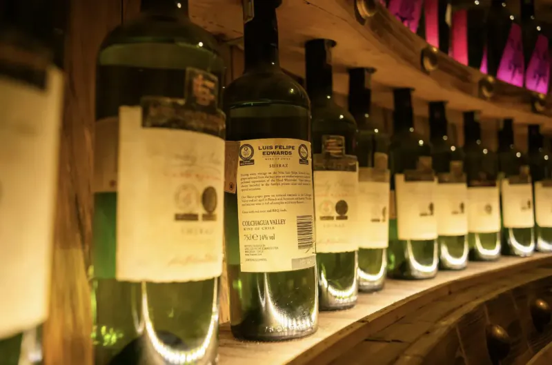Close-up of green wine bottles with labels stored on wooden shelves in a dimly lit cellar, warm lighting.