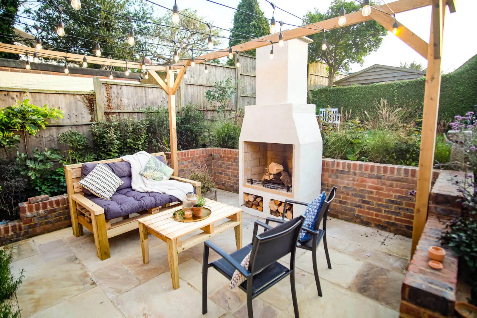 Schiedel Isokern Volcanic white garden fireplace with burning logs, cozy wooden bench, chairs, low table, string lights, and plants on patio.