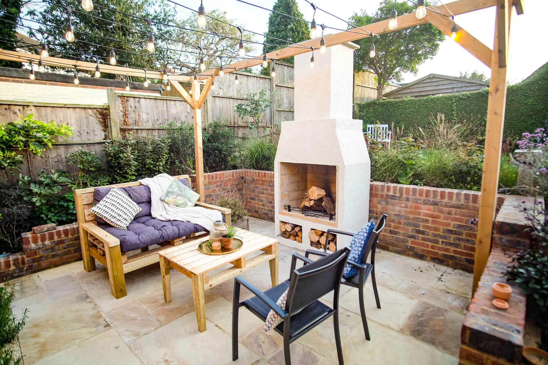 Schiedel Isokern Volcanic white garden fireplace with burning logs, cozy wooden bench, chairs, low table, string lights, and plants on patio.