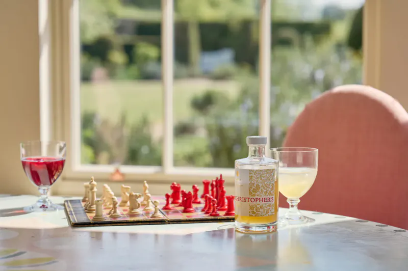 Chessboard with red and white pieces on table beside Christopher’s Cordials bottle, red wine, and yellow cordial glasses, sunlit window view.