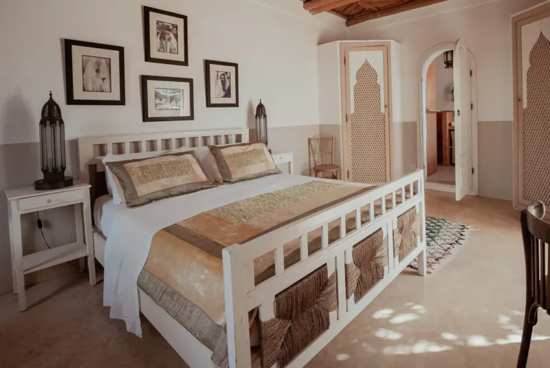 Moroccan-style bedroom in Essaouira retreat with white wood king bed, beige linens, lanterns, arched doors, and framed art.
