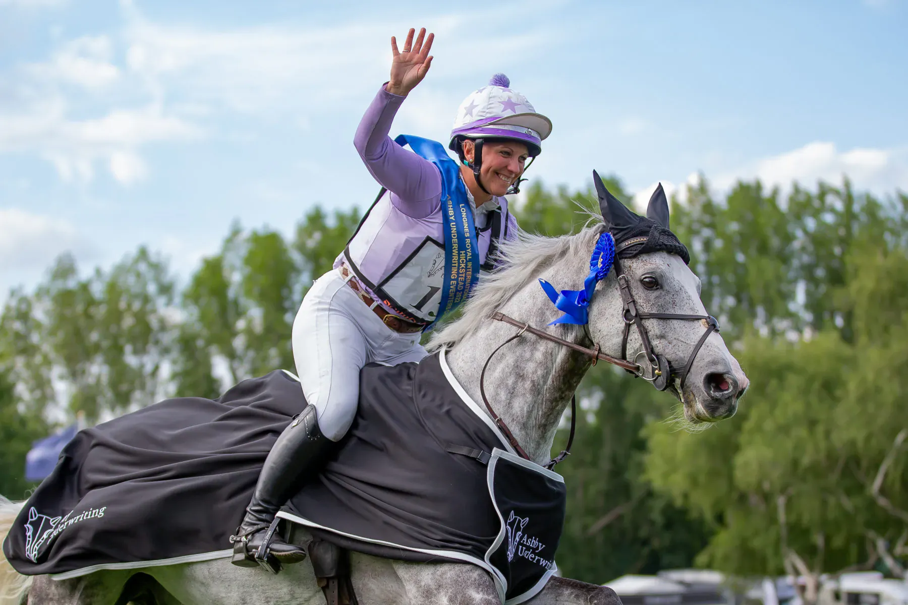 Female rider in purple vest and helmet waves atop gray horse with black blanket at Hickstead Derby, trees backdrop