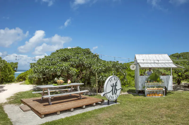 White 'HF' sign outside wooden stand with fruits on table, picnic bench, lush greenery, beach path under blue sky