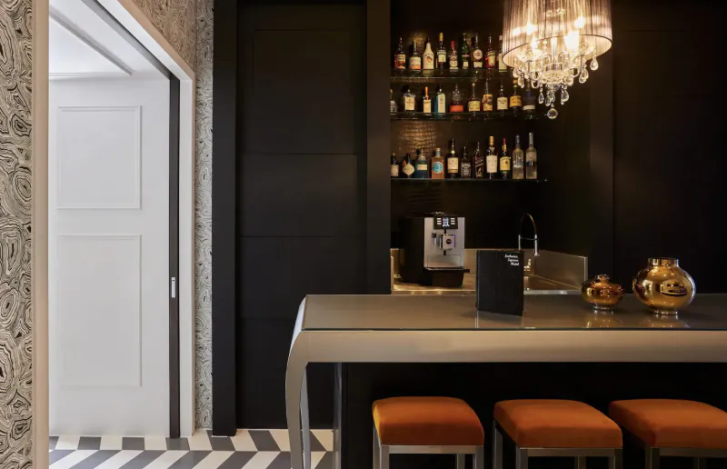 Elegant boutique hotel bar with black walls, lit chandelier, liquor shelves, coffee machine, sleek counter, and orange stools