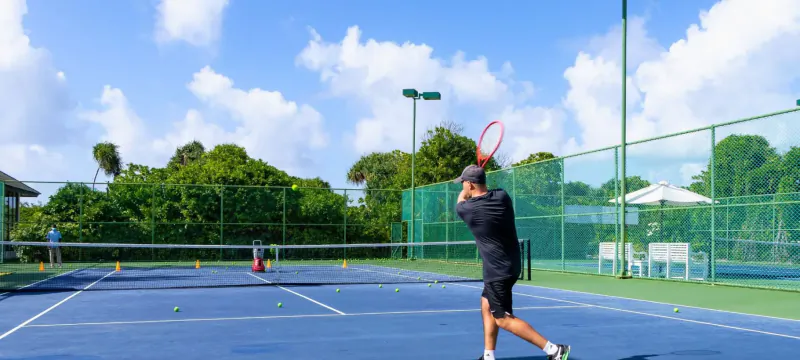 Man in black swinging red tennis racket on outdoor court at Hideaway Beach Resort, Maldives, lush greenery backdrop.