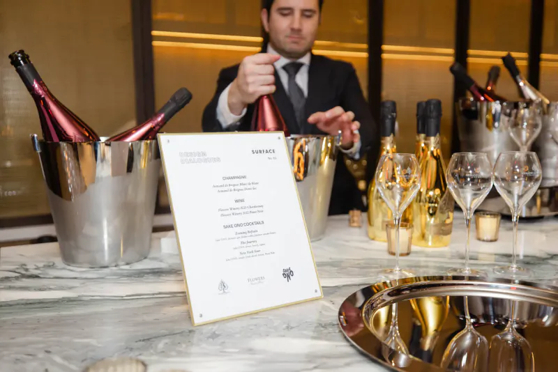 Man in suit pouring champagne beside ice buckets of bottles and menu at upscale New York industry event