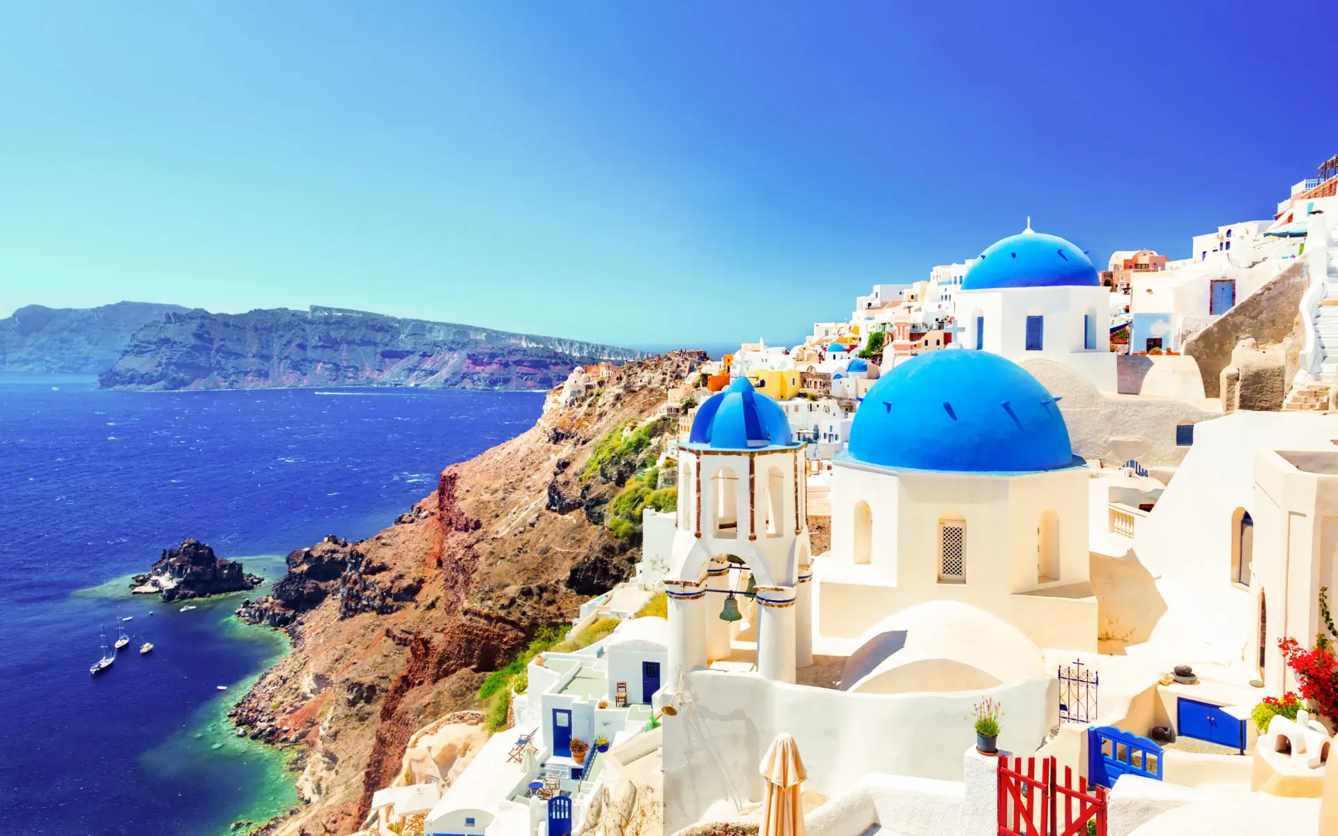 Oia, Santorini: white buildings with blue domes cascading down cliffs to turquoise Aegean Sea under clear blue sky