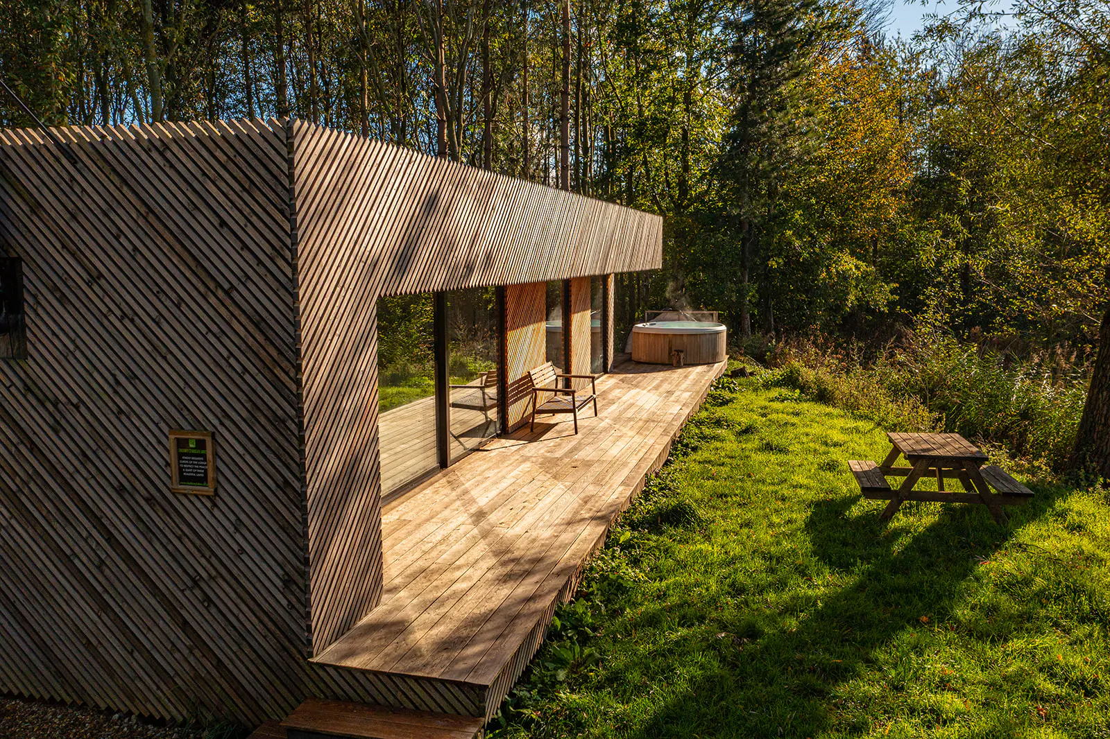 Modern wooden glamping cabin with deck, hot tub, chairs, picnic table in autumn forest, Essex.