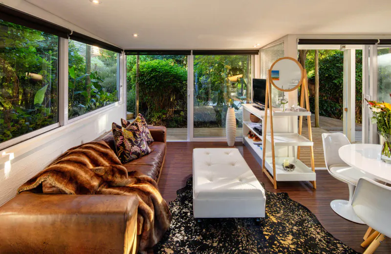 Bright modern living room at Spanish Farm Guest Lodge with brown leather sofa, white ottoman, round mirror, plants, and garden view.