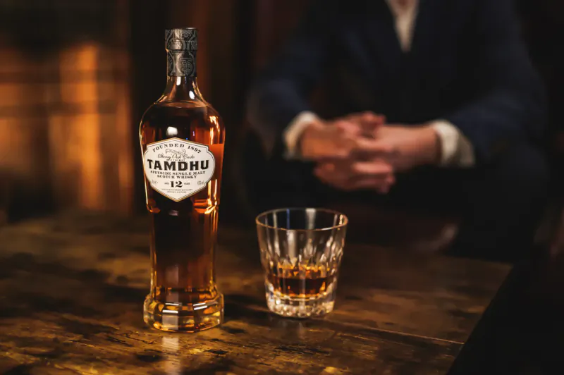 Tamdhu whisky bottle and glass of amber whisky on wooden table, blurred man in background.