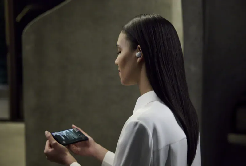Asian woman with long black hair wearing wireless earbuds, holding smartphone, side profile in dim setting