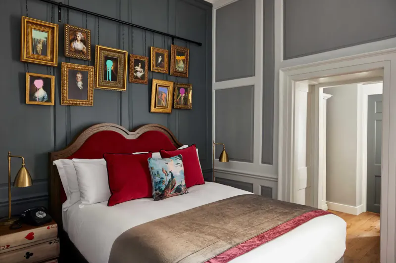 Luxurious Hotel Indigo Bath bedroom with red velvet bed, gold-framed portraits on gray wall, and arched doorway.