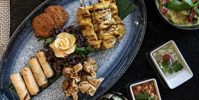 Thai platter on blue plate: chicken satay skewers, spring rolls, dumplings, fish cakes, garnished with rose, with green curry and sauces