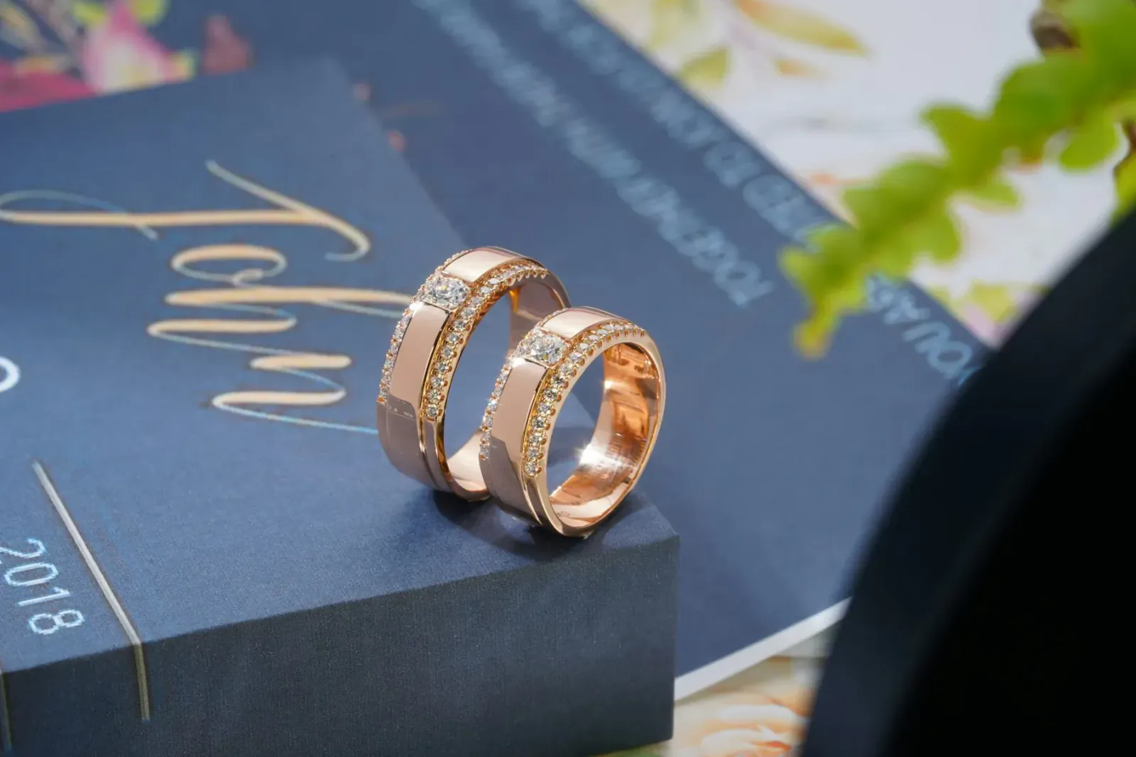 Pair of rose gold diamond wedding rings on black box with blue 'John 2018' invitation and flowers