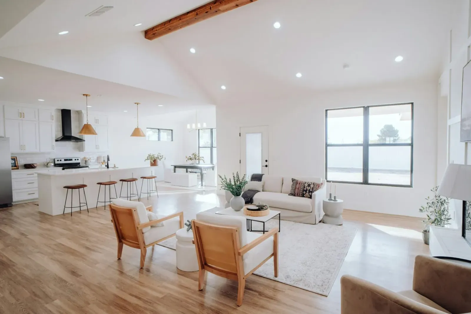 Modern open-plan living room with white kitchen, wooden beams, plush sofa, chairs, plants, and large windows