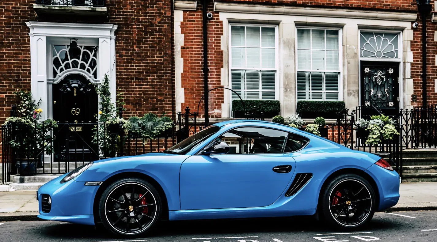 Bright blue Porsche 718 Cayman sports car parked on street in front of elegant brick townhouses with wrought-iron fences.
