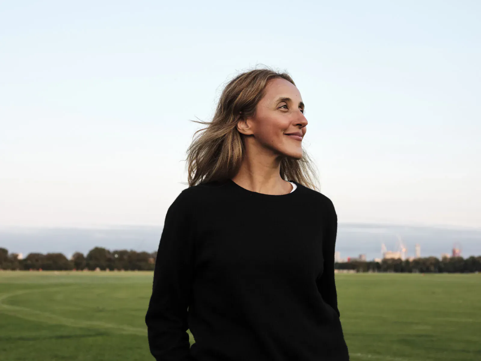 Blonde woman smiling sideways in black sweater, windblown hair, green field and city skyline backdrop