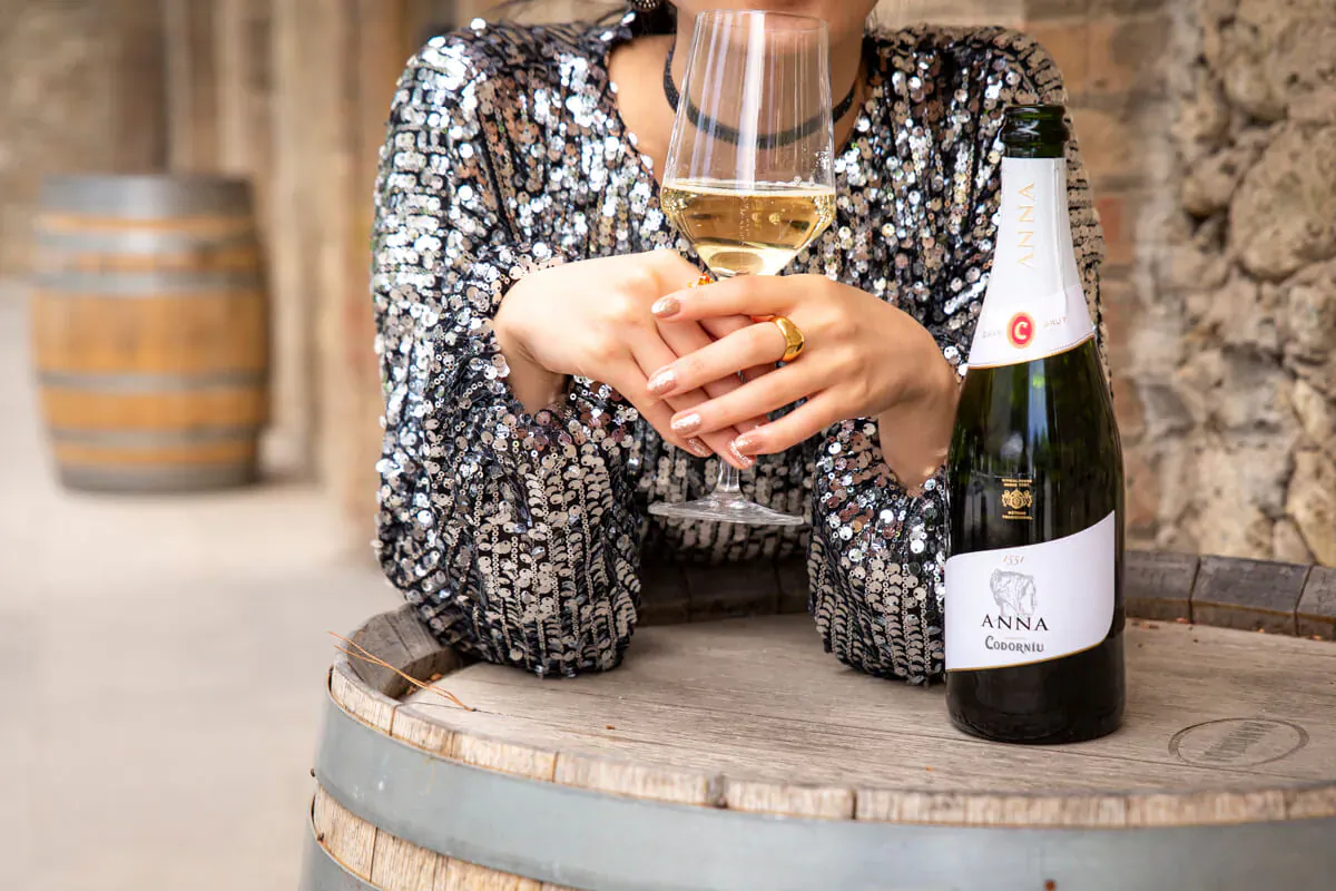 Woman in sparkly dress holds glass of white wine, Codorníu Cava bottle on barrel in stone courtyard