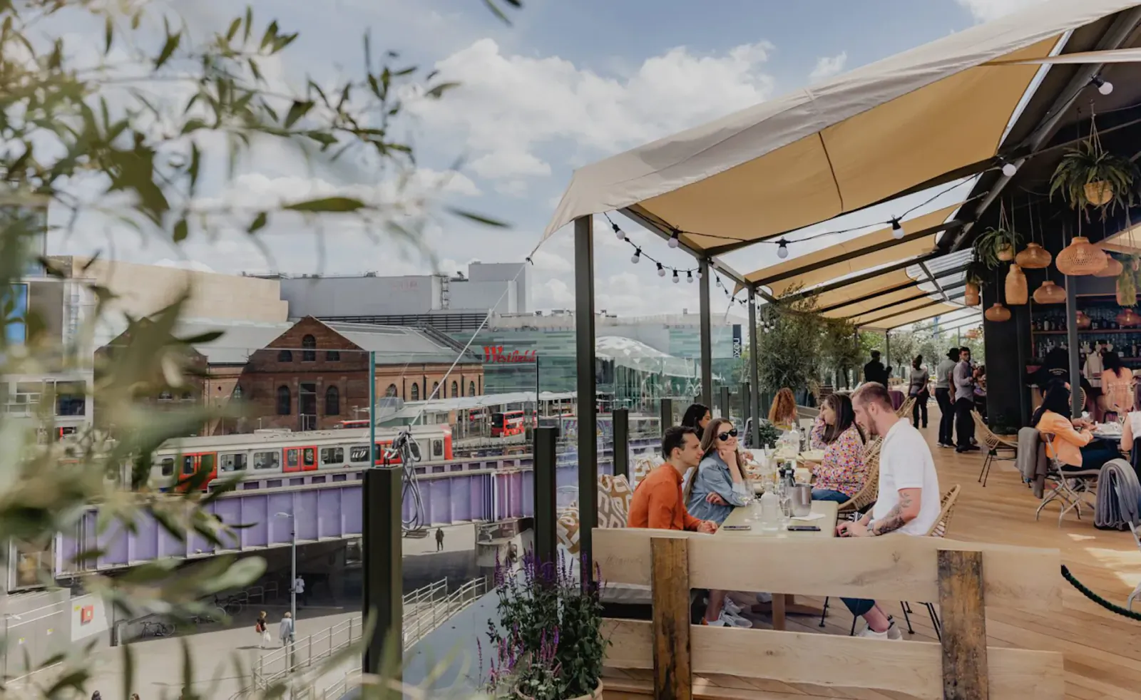 People dining outdoors under beige canopy at The Broadcaster, London, with panoramic canal, bridge, and city views.