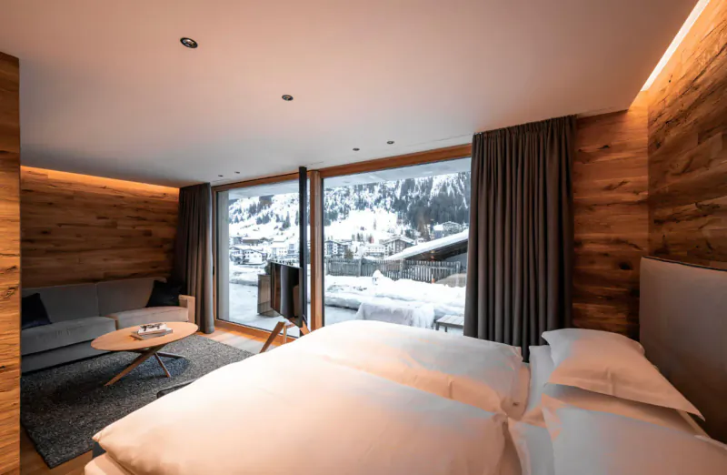 Cozy wooden luxury chalet bedroom with king bed, sofa, snowy alpine mountain view through large windows.