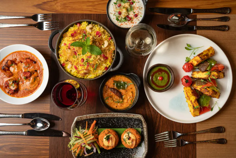 Indian thali on wooden table: butter chicken, biryani, paneer tikka, naan, chutneys, wine
