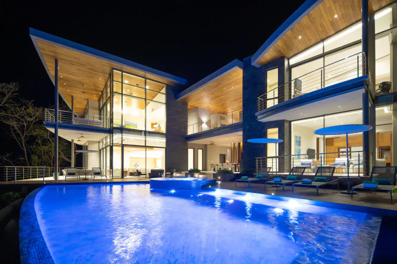 Modern luxury bespoke home at night with glowing blue infinity pool, lounge chairs, and ocean views.