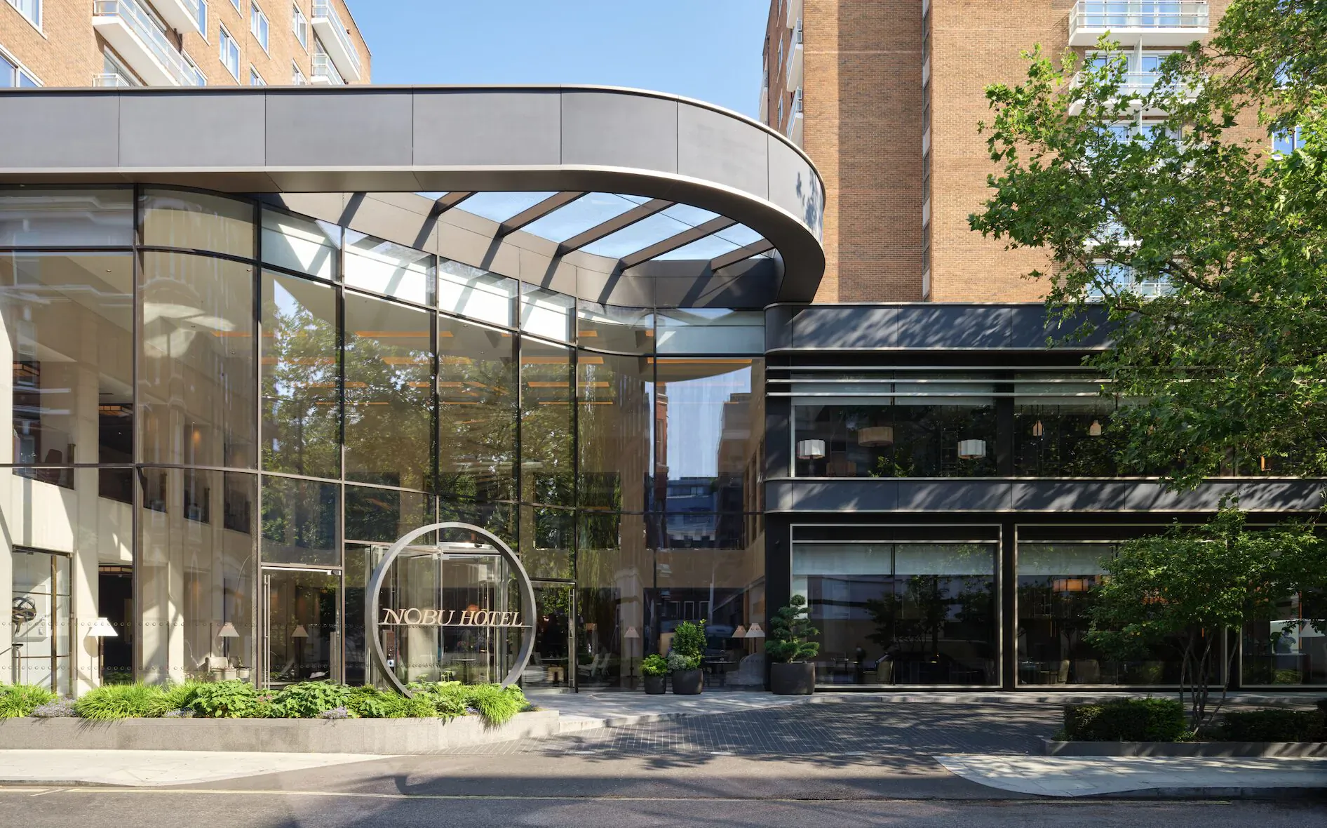 Nobu Hotel London Portman Square entrance with glass canopy, circular sign, brick buildings, trees, and plaza in Marylebone.