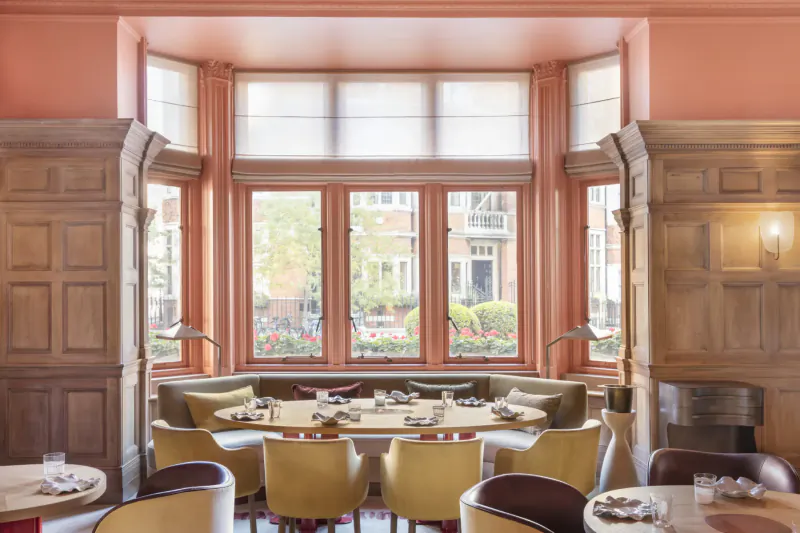Elegant dining room at Hélène Darroze at The Connaught with bay window view of garden, round table, cushioned chairs.