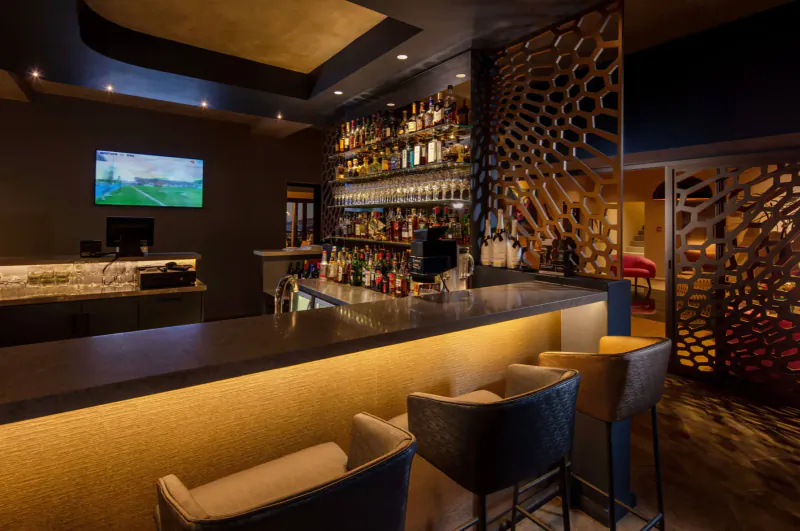 Modern hotel bar with wooden lattice dividers, stocked shelves of liquor bottles, backlit counter, bar stools, and golf TV screen.
