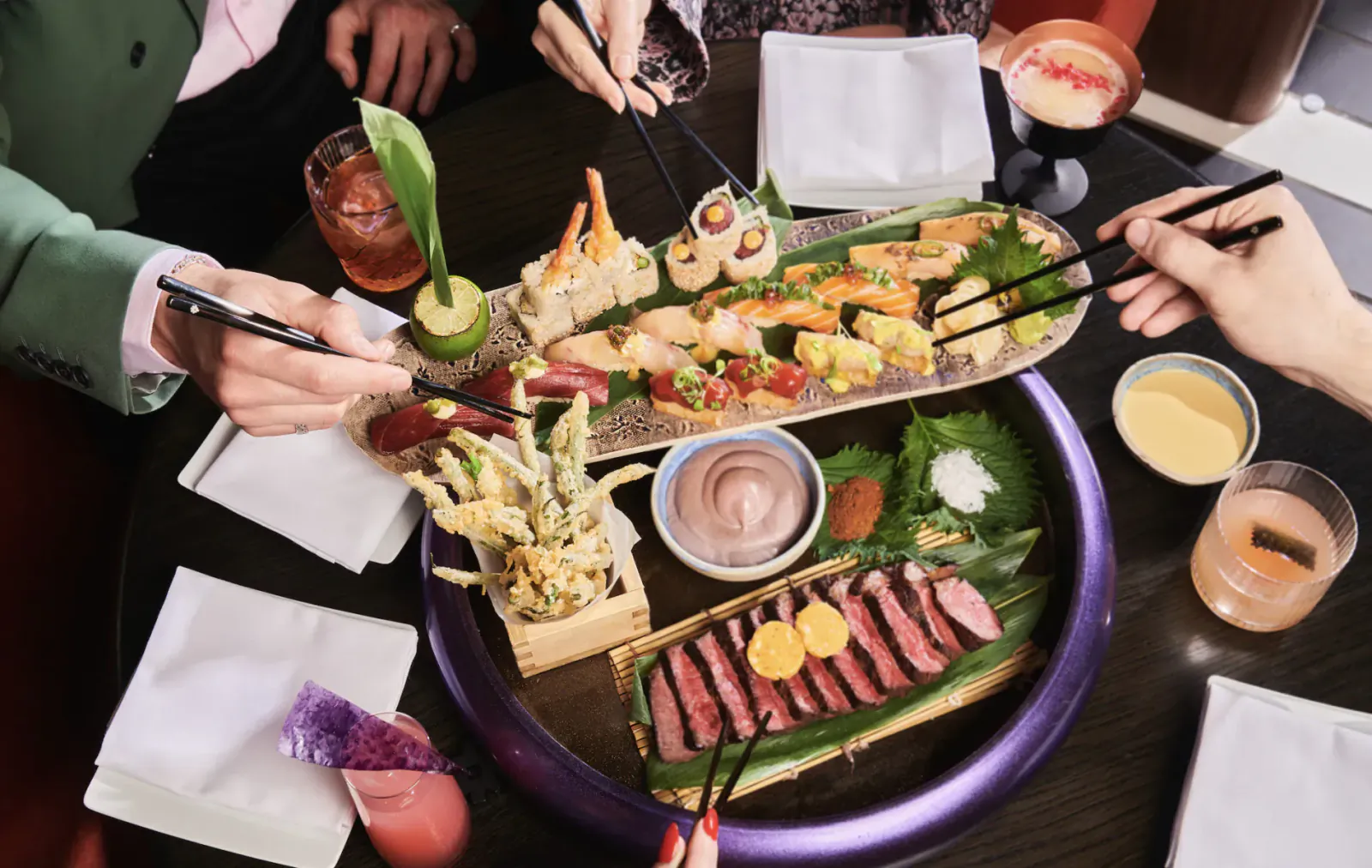 Overhead view of Chotto Matte Sunday set menu: sushi, sashimi, wagyu, tempura on purple platter, hands using chopsticks, cocktails.
