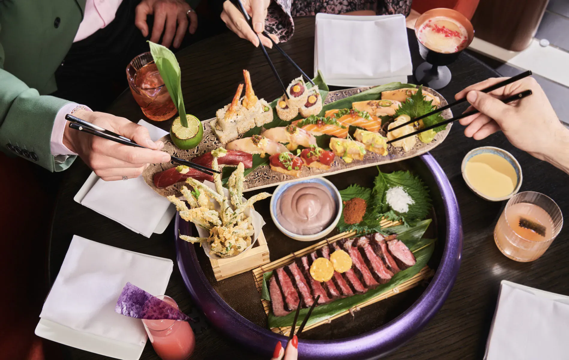 Overhead view of Chotto Matte Sunday set menu: sushi, sashimi, wagyu, tempura on purple platter, hands using chopsticks, cocktails.