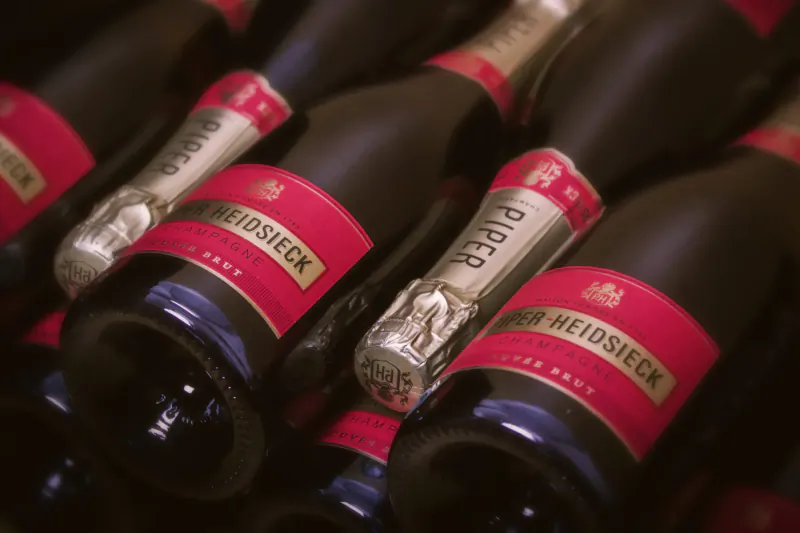 Close-up of Piper-Heidsieck champagne bottles with red labels in a dark rack, premium holiday indulgence.