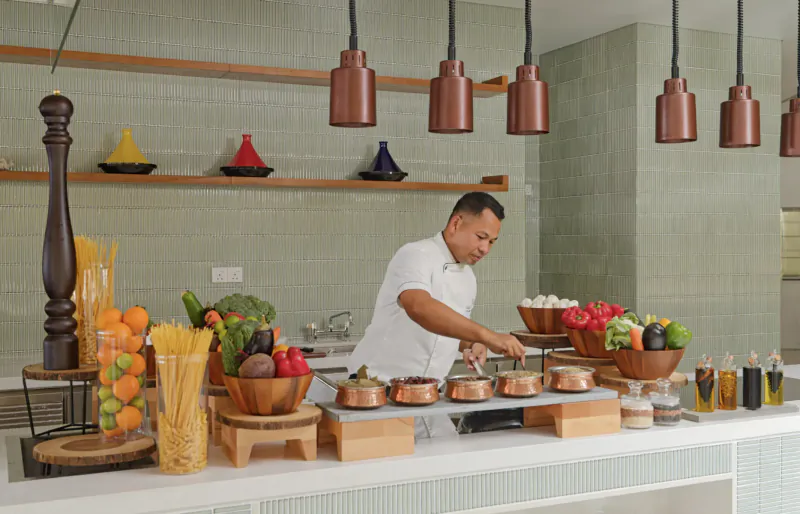 Chef in white uniform preparing food on kitchen counter with colorful Moroccan lamps, fresh vegetables, pasta, and pots.