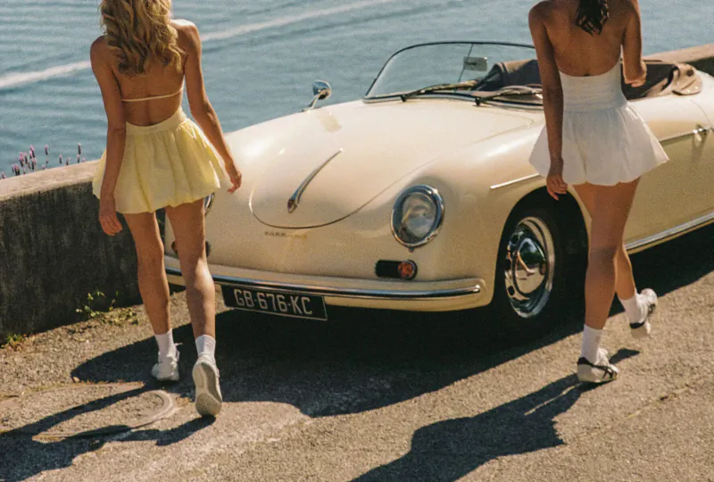 Two women in yellow and white mini skirts walk beside a cream Porsche 356 sports car by the sea, license plate GB51K. Riviera floral fashion.