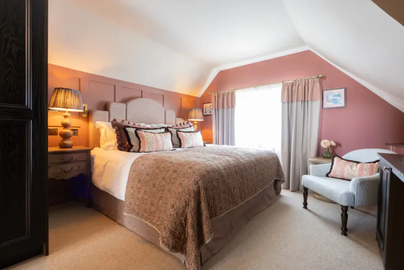 Cozy attic bedroom in The George & Dragon with king bed, pink walls, lamp, armchair, and window.