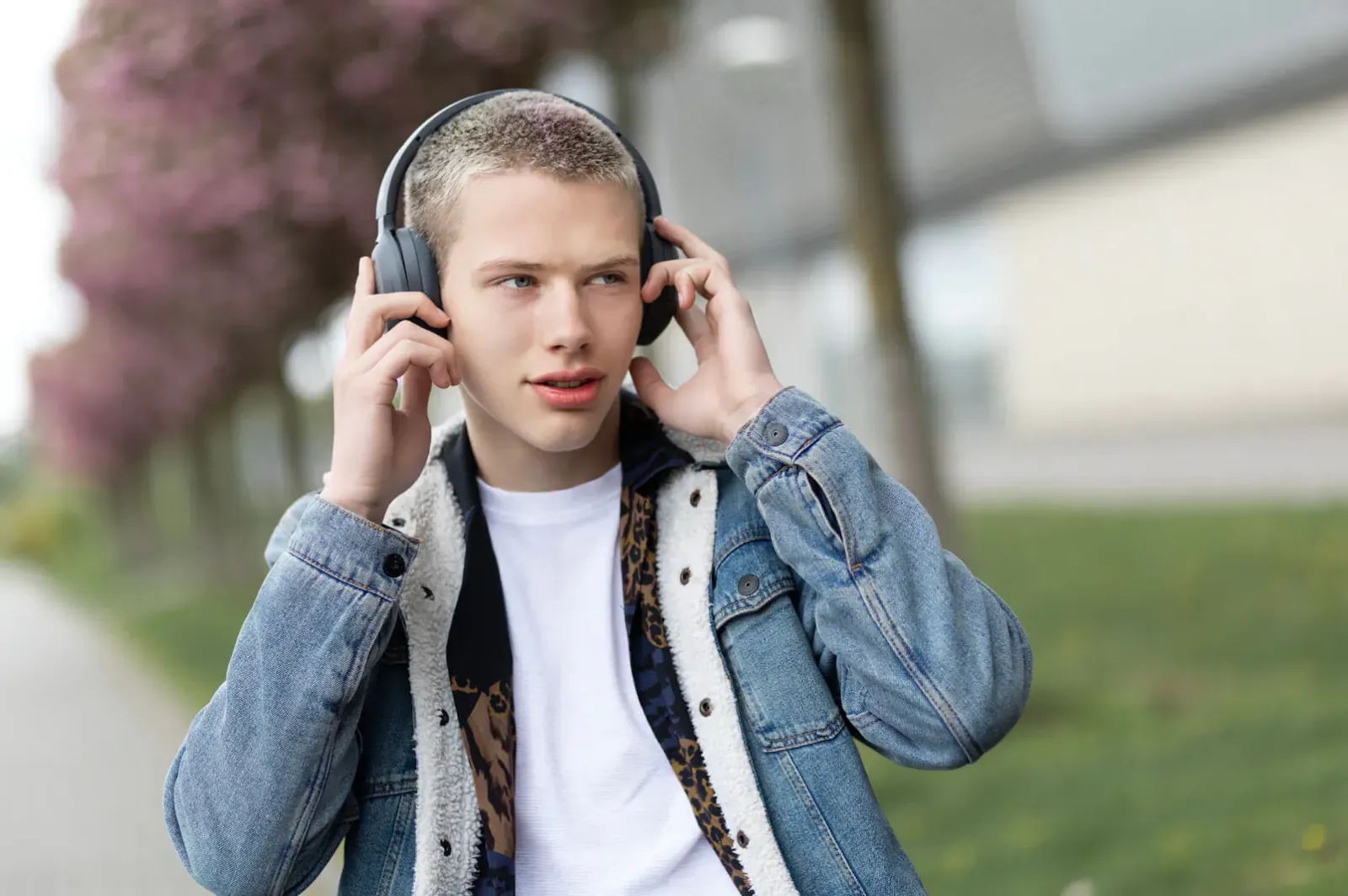 Young man with buzz cut wearing Edifier W820NB headphones, adjusting them outdoors by cherry trees and modern building