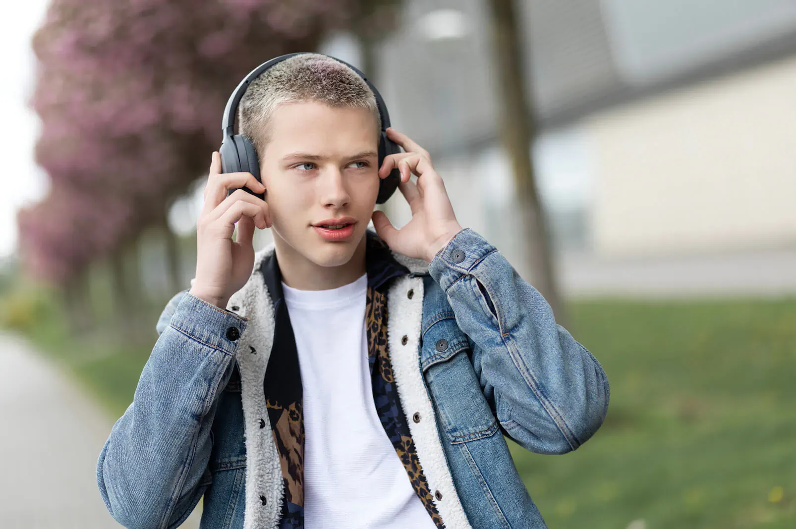 Young man with buzz cut wearing Edifier W820NB headphones, adjusting them outdoors by cherry trees and modern building