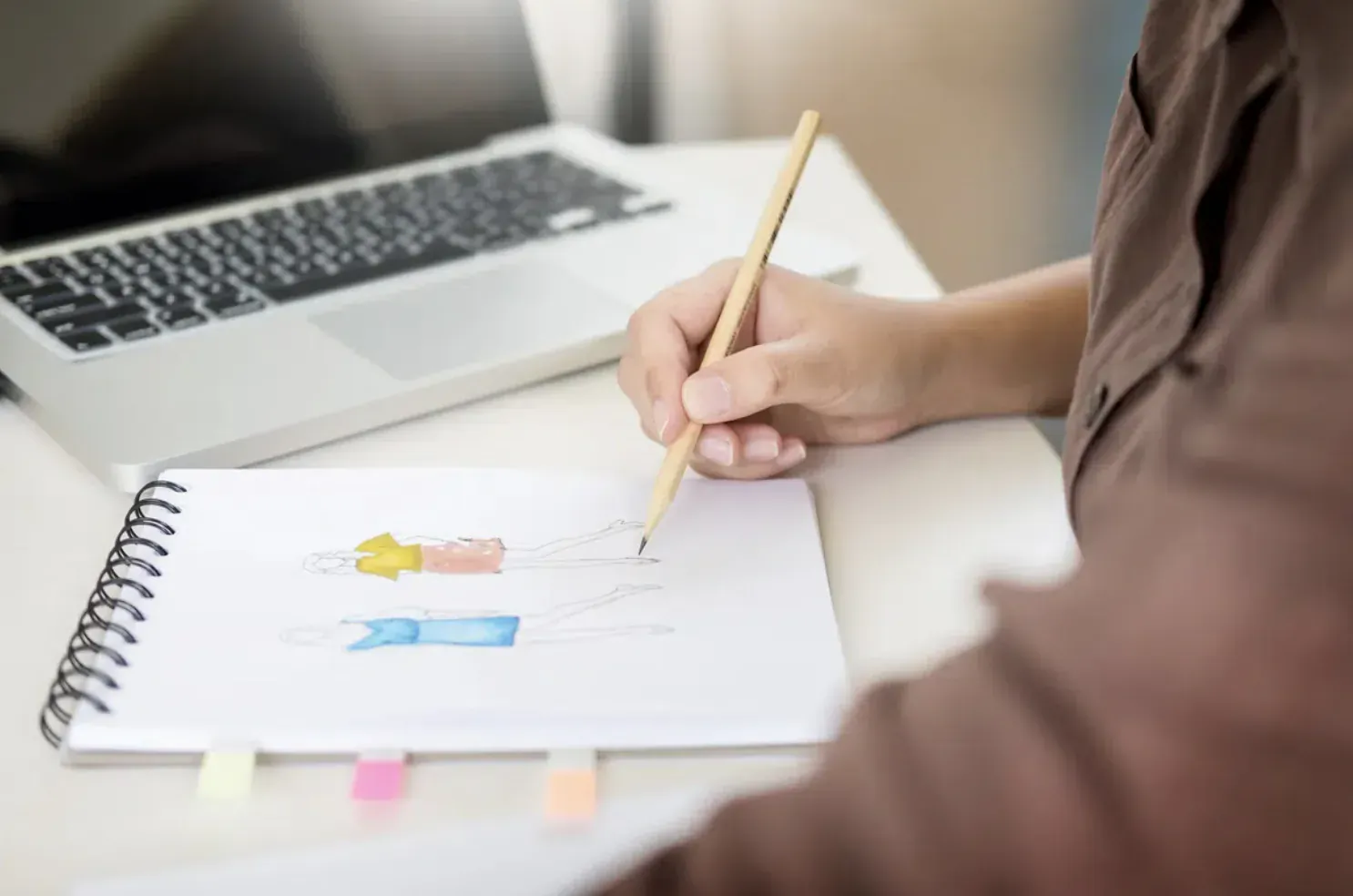 Person sketching colorful dress designs in notebook beside laptop on desk