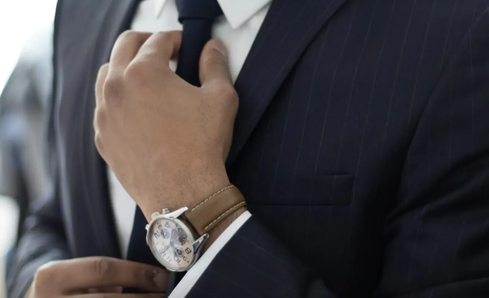 Man in suit adjusting navy tie, wearing white shirt and brown leather watch