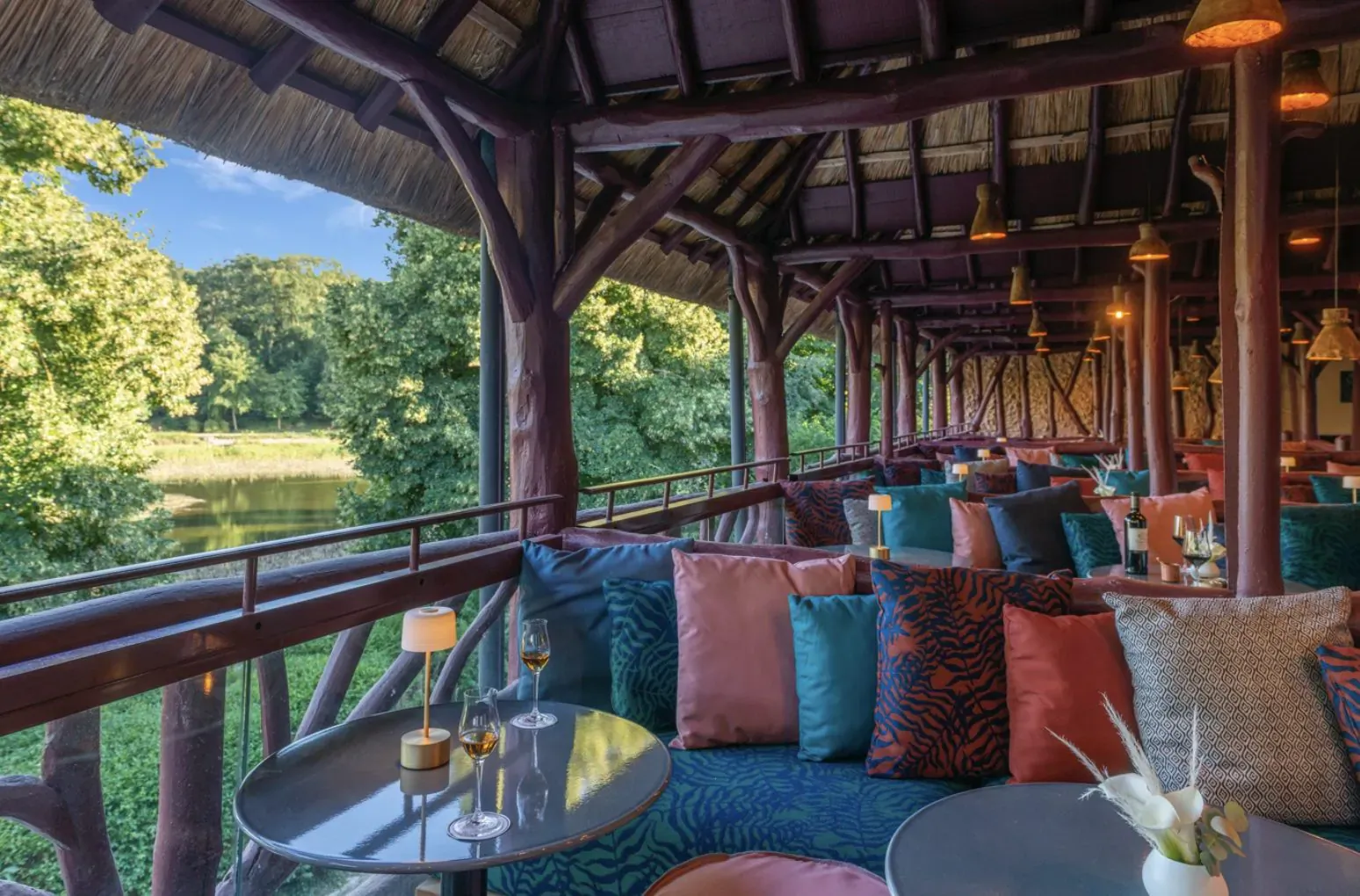Luxurious outdoor terrace at Les Étangs de Corot Paris with colorful cushions, low tables, and lake view through lush trees.